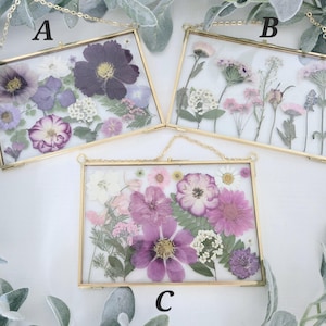 May include: Three gold framed pictures with pressed flowers. The frames are hanging from a chain. The pictures are labeled A, B, and C. The flowers are in shades of purple, pink, and white.