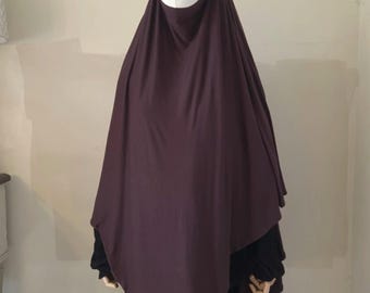 May include: A dark brown, solid-colored, one-piece hijab that drapes over the head and shoulders, extending down to the chest area. The fabric is opaque and smooth, worn over a black long-sleeved garment on a mannequin with a white head.