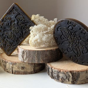Può includere: Tre saponette decorative con intricati disegni floreali, esposte su rotonde di legno. I saponi sono di colore marrone scuro e verde con una superficie testurizzata.