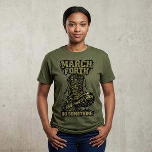 May include: An olive green t-shirt featuring the text "MARCH FORTH" above a graphic of a combat boot. Below the boot, the text reads "DO SOMETHING". The shirt is worn by a person with dark hair and blue jeans.