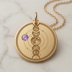 May include: Gold-colored pendant necklace featuring a detailed engraving of the moon phases. A small, round, purple gemstone is set on the pendant. The necklace has a delicate gold chain and a circular pendant design.