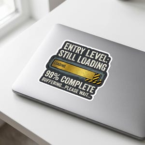 May include: A white sticker on a laptop with the text "ENTRY LEVEL: STILL LOADING" above a loading bar graphic. Below the graphic is the text "99% COMPLETE BUFFERING...PLEASE WAIT..."