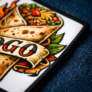 May include: Embroidered patch featuring a detailed illustration of a burrito with flames and a banner that reads "GO". The patch has a black border and is set against a blue denim background.