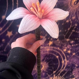 May include: A handmade, large pink and white flower with a green stem. The flower has multiple petals with a textured surface and a light pink gradient. The center of the flower has orange stamens. The stem is covered in green velvet.