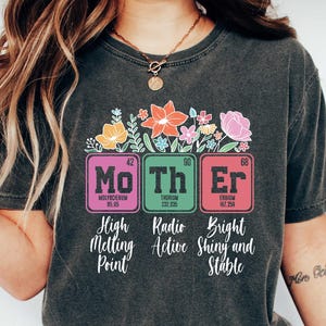 May include: A dark gray t-shirt with a graphic of the periodic table elements Mo, Th, and Er, spelling out the word "Mother". The graphic is surrounded by colorful flowers. The text below the graphic reads "High Melting Point", "Radio Active", and "Bright Shiny and Stable".