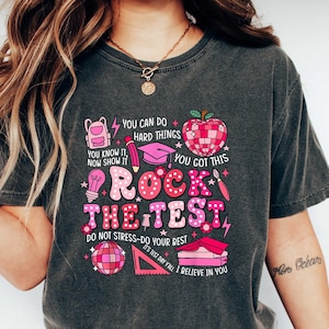 May include: A dark gray t-shirt with a pink and white graphic design that says "You can do hard things. You know it, now show it. You got this. Rock the test. Do not stress-do your best. It's test day y'all. I believe in you."