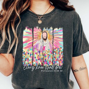 Puede incluir: Una camiseta gris oscuro con un gr&aacute;fico colorido de Jes&uacute;s rodeado de una multitud de personas. El gr&aacute;fico tiene un efecto de salpicaduras de pintura e incluye el texto "Every knee shall bow" y "Philippians 2:10-11".