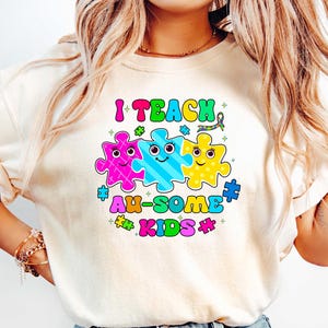 May include: A white t-shirt with the text "I Teach Au-Some Kids" in rainbow colors. The text is surrounded by three puzzle pieces with smiling faces.