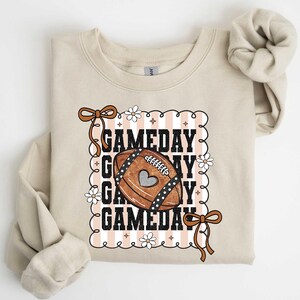 May include: A beige sweatshirt with a football graphic that says "GAMEDAY" in a decorative frame. The football is brown with a heart and white stitching. The sleeves are rolled up.