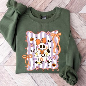 May include: Olive green sweatshirt featuring a Halloween-themed graphic. The graphic includes a ghost reading a book, surrounded by flowers, pumpkins, and bats, set against a striped background with orange bows.