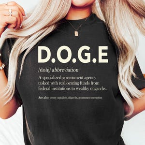 May include: Black t-shirt with the white text "D.O.G.E" and a definition. The definition describes a specialized government agency. The shirt also includes the pronunciation and related terms.