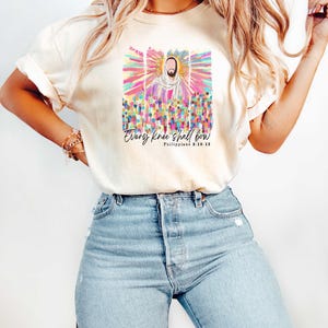 Puede incluir: Una camiseta blanca con un gr&aacute;fico colorido de Jes&uacute;s con el texto "Every knee shall bow" y "Philippians 2:10-13" impreso en la parte delantera.