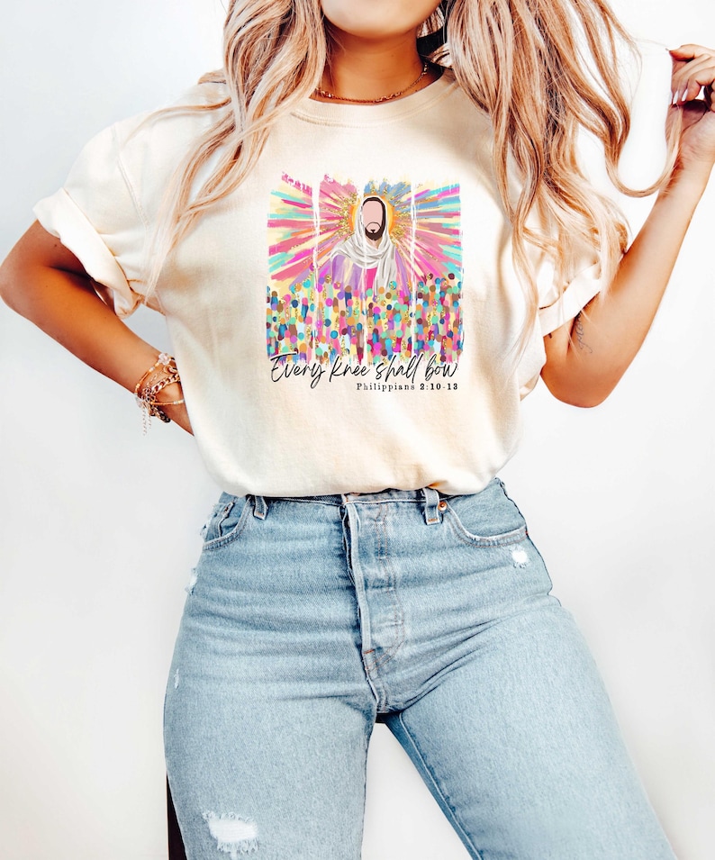 Puede incluir: Una camiseta blanca con un gr&aacute;fico colorido de Jes&uacute;s con el texto "Every knee shall bow" y "Philippians 2:10-13" impreso en la parte delantera.