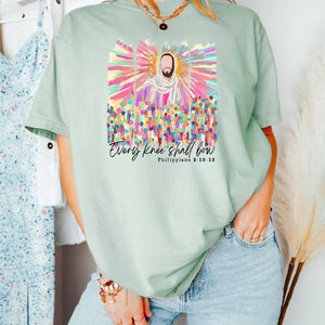 Puede incluir: Una camiseta verde claro con un gr&aacute;fico colorido de una persona rodeada de una multitud. El texto "Every knee shall bow" y "Philippians 2:10-13" est&aacute; impreso debajo del gr&aacute;fico.