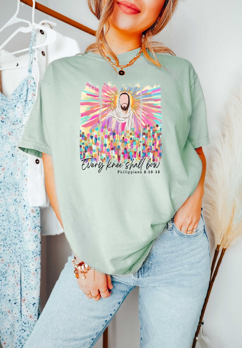 Puede incluir: Una camiseta verde claro con un gr&aacute;fico colorido de una persona rodeada de una multitud. El texto "Every knee shall bow" y "Philippians 2:10-13" est&aacute; impreso debajo del gr&aacute;fico.
