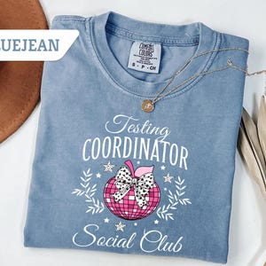 May include: Bluejean colored t-shirt with the words "Testing Coordinator Social Club" in white script. A pink disco ball apple with a black and white bow is in the center. A gold necklace and a brown hat are also in the image.
