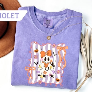 May include: Violet-colored t-shirt with a graphic of a ghost reading a book, adorned with flowers, pumpkins, and bows. The ghost is set against a striped background. A gold necklace is draped on the shirt. The word "VIOLET" is in the upper left corner.