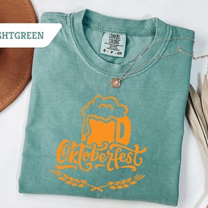 May include: Light green t-shirt with an orange Oktoberfest beer stein graphic. The shirt has a crew neck and short sleeves. A silver necklace with a pendant is draped on the shirt. The word "LIGHTGREEN" is in a white box in the upper left corner.