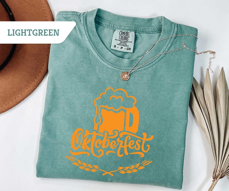 May include: Light green t-shirt with an orange Oktoberfest beer stein graphic. The shirt has a crew neck and short sleeves. A silver necklace with a pendant is draped on the shirt. The word "LIGHTGREEN" is in a white box in the upper left corner.