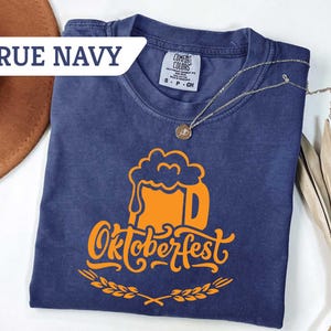 May include: A navy blue t-shirt with an orange Oktoberfest beer mug graphic. The text "Oktoberfest" is below the mug, with a wheat design. The shirt has a tag that says "Comfort Colors". A gold necklace is draped across the shirt.