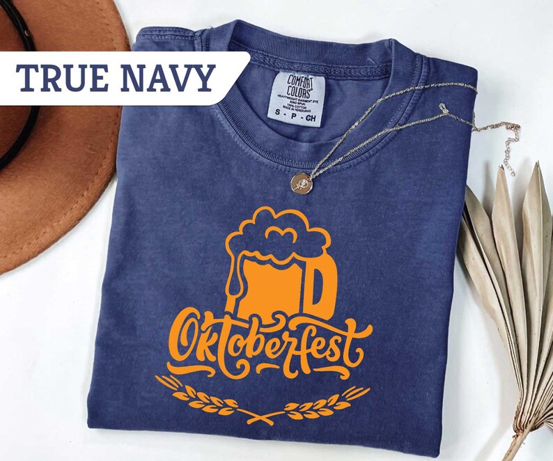 May include: A navy blue t-shirt with an orange Oktoberfest beer mug graphic. The text "Oktoberfest" is below the mug, with a wheat design. The shirt has a tag that says "Comfort Colors". A gold necklace is draped across the shirt.