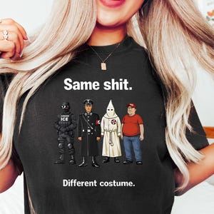 May include: Black t-shirt with the text "Same shit. Different costume." and illustrations of four figures in different uniforms and attire. The figures include a person in a black uniform with "ICE" on it, a person in a black uniform, a person in a white robe and hood, and a person in a red shirt and a red cap.
