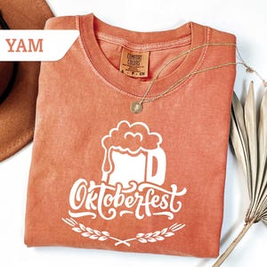 May include: A coral-colored t-shirt with a white Oktoberfest beer stein graphic. The design includes the word "Oktoberfest" in a decorative font, with a wheat sprig border. A gold necklace and a hat are also in the image.