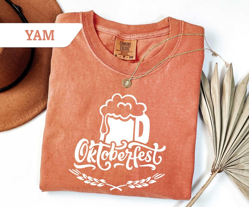 May include: A coral-colored t-shirt with a white Oktoberfest beer stein graphic. The design includes the word "Oktoberfest" in a decorative font, with a wheat sprig border. A gold necklace and a hat are also in the image.