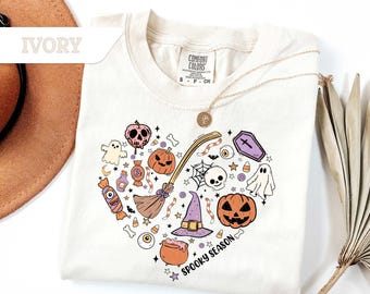 May include: Ivory-colored t-shirt with a heart-shaped Halloween design. The design includes a skull, pumpkins, ghosts, a witch's hat, and the words "Spooky Season". A gold necklace is draped over the shirt.
