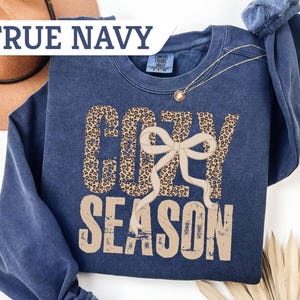 May include: A navy blue crewneck sweatshirt featuring the text COZY SEASON in a distressed font. The word COZY is filled with a brown and black leopard print pattern, and a beige ribbon bow graphic is layered over the letters. The word SEASON is solid beige.