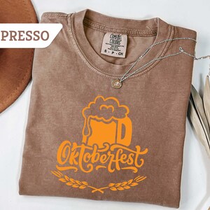 May include: Espresso-colored t-shirt with an orange Oktoberfest beer stein graphic. The shirt has a crew neck and short sleeves. A silver necklace with a pendant is draped across the shirt. A brown hat and dried plant are in the background.