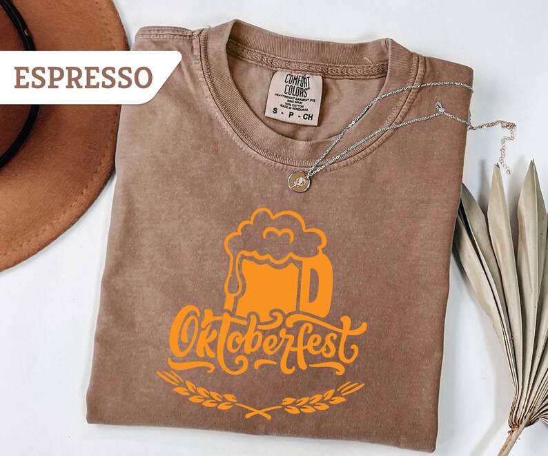 May include: Espresso-colored t-shirt with an orange Oktoberfest beer stein graphic. The shirt has a crew neck and short sleeves. A silver necklace with a pendant is draped across the shirt. A brown hat and dried plant are in the background.