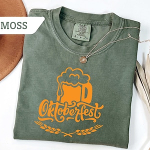 May include: Moss green t-shirt with an orange Oktoberfest beer mug graphic. The shirt has a crew neck and short sleeves. A gold necklace with a pendant is draped across the shirt. A brown hat is visible in the upper left corner.