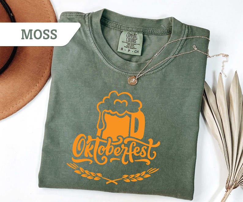 May include: Moss green t-shirt with an orange Oktoberfest beer mug graphic. The shirt has a crew neck and short sleeves. A gold necklace with a pendant is draped across the shirt. A brown hat is visible in the upper left corner.