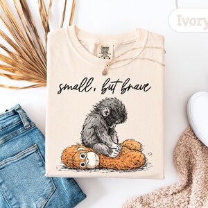 May include: Ivory-colored t-shirt featuring a graphic of a small, gray monkey hugging a brown stuffed animal. The text "small, but brave" is printed above the monkey. The shirt is laid flat with a pair of blue jeans and white shoes.