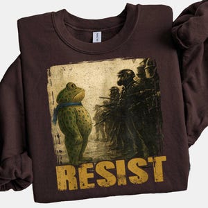 May include: A dark brown sweatshirt featuring a distressed graphic of a frog wearing a blue scarf facing a line of soldiers. The word "RESIST" is printed in yellow block letters below the image. The sweatshirt is made of a soft material.
