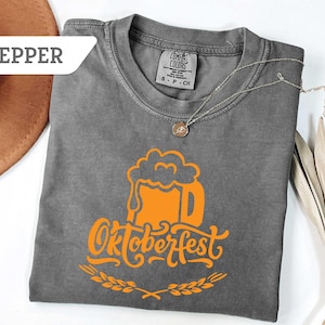 May include: A gray Comfort Colors t-shirt with an orange Oktoberfest beer mug graphic. The design includes the word "Oktoberfest" in a stylized font, with a wheat sprig at the bottom. A silver necklace with a pendant is draped on the shirt.
