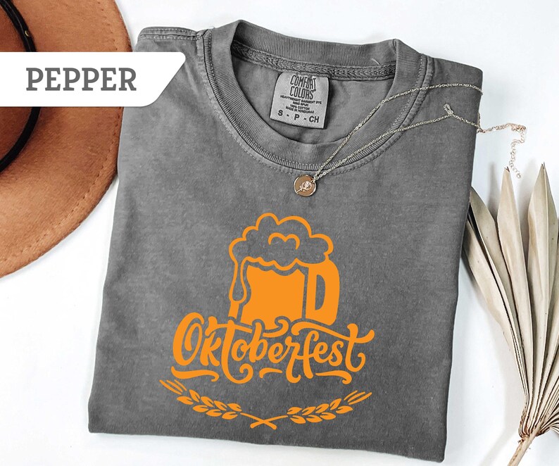 May include: A gray Comfort Colors t-shirt with an orange Oktoberfest beer mug graphic. The design includes the word "Oktoberfest" in a stylized font, with a wheat sprig at the bottom. A silver necklace with a pendant is draped on the shirt.