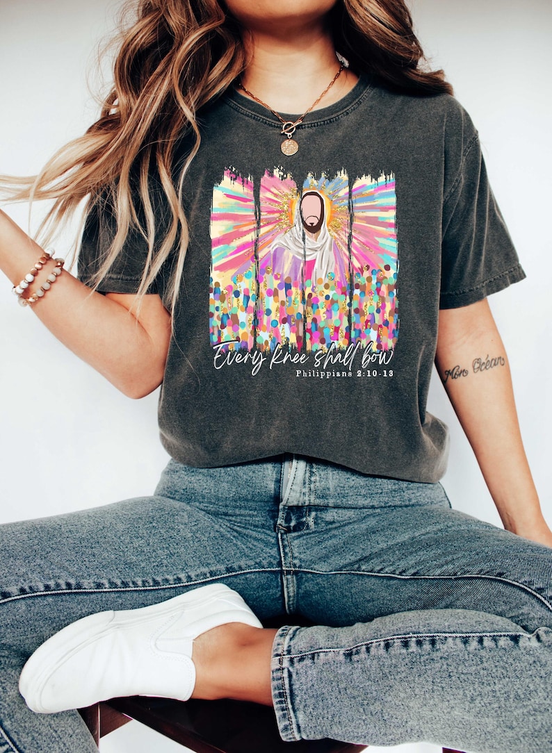 Puede incluir: Una camiseta gris oscuro con un gr&aacute;fico colorido de Jes&uacute;s rodeado de una multitud de personas. El gr&aacute;fico tiene un efecto de salpicaduras de pintura e incluye el texto "Every knee shall bow" y "Philippians 2:10-11".
