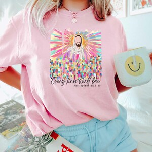 Puede incluir: Camiseta rosa con un gr&aacute;fico colorido de Jes&uacute;s rodeado de personas. El gr&aacute;fico incluye el texto "Every knee shall bow" y "Philippians 2:10-13".