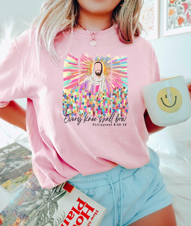 Puede incluir: Camiseta rosa con un gr&aacute;fico colorido de Jes&uacute;s rodeado de personas. El gr&aacute;fico incluye el texto "Every knee shall bow" y "Philippians 2:10-13".