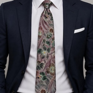 May include: A necktie featuring a muted mauve and grey background with a detailed floral pattern in shades of dark green, brown, and yellow. The tie is worn with a crisp white dress shirt, a navy blue blazer, and a white pocket square.