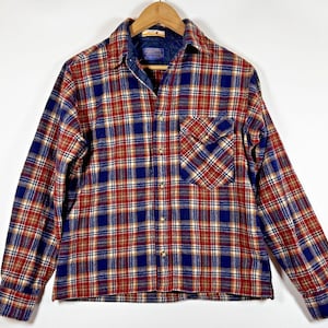 May include: A long-sleeved, button-down flannel shirt in a classic plaid pattern. The shirt features a combination of red, blue, and tan colors. It has a chest pocket and a collar with a blue lining. The shirt is hanging on a wooden hanger.