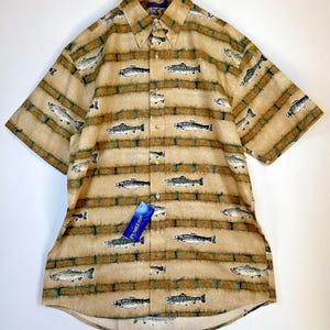 May include: Tan short-sleeve button-down shirt with a fish pattern. The shirt features a repeating pattern of fish in shades of gray and green, set against a background of horizontal tan and brown stripes. A blue tag with the brand name is attached.