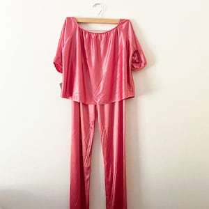 Puede incluir: Un conjunto de pijama de satén rosa de dos piezas. La parte superior tiene un cuello ancho y mangas cortas. Los pantalones son largos y de corte recto. El conjunto está colgado en una percha de madera contra una pared blanca. La tela tiene un ligero brillo.