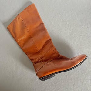 Puede incluir: Una bota alta de cuero naranja. La bota tiene una punta redondeada y un tacón bajo. El cuero parece desgastado y arrugado, lo que sugiere un estilo vintage. La bota está en ángulo, mostrando su perfil lateral sobre un fondo neutro.