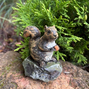 Puede incluir: Figurilla detallada de una ardilla, posada sobre una pequeña roca, sosteniendo una nuez. La ardilla tiene pelaje marrón y gris con una cola tupida. La figurilla está colocada sobre un fondo de follaje verde y una piedra marrón rojiza.