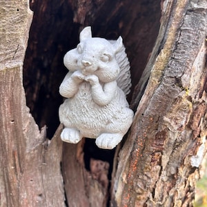 Puede incluir: Una figurita de ardilla de hormigón gris con cola tupida, sentada dentro de un tronco de árbol con corteza marrón.