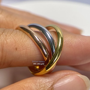 Könnte beinhalten: Ein dreifarbiges Ring mit drei ineinander verschlungenen Bändern in Gold, Roségold und Silber. Der Ring wird an einem Finger gezeigt, mit dem Text "D Gold District 18K solid gold" sichtbar.