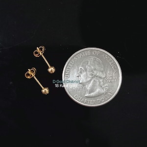 May include: Two small, gold-coloured stud earrings with a ball design are displayed next to a quarter. The earrings are on a black surface. The text "D Gold District 18 Karat gold" is visible.
